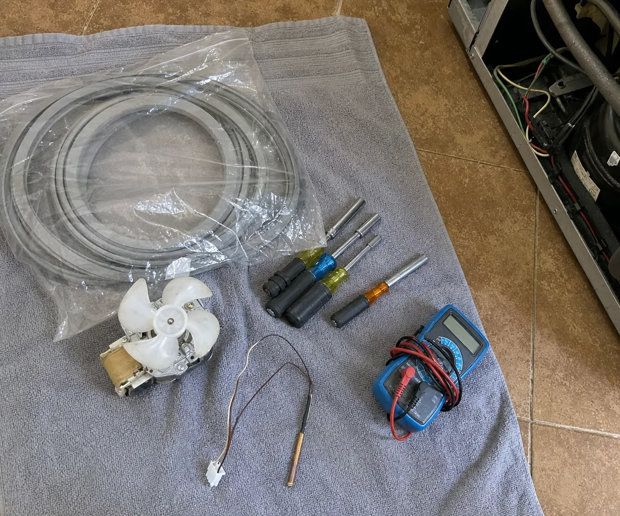 Mirrored view of a repair kit on a towel with a water supply line, screwdrivers and test leads, appliance chassis open in the frame