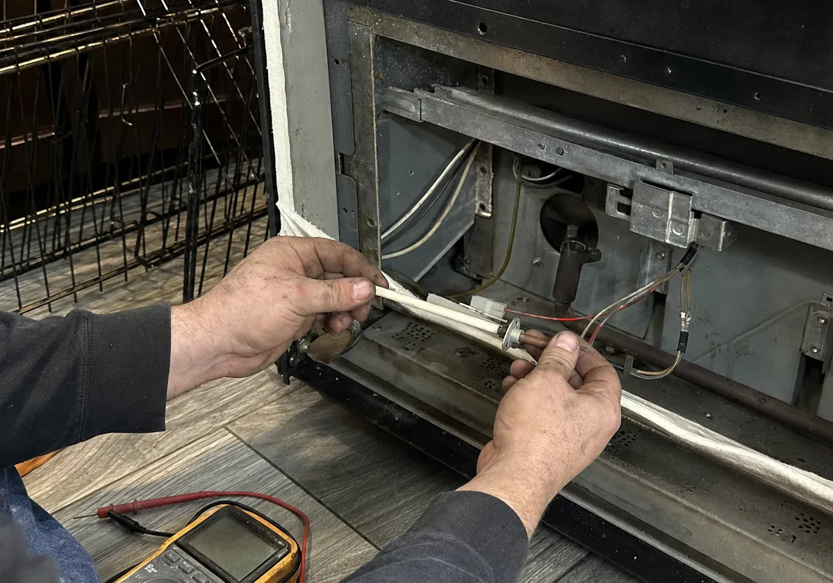 Close view of hands at an exposed burner tube and igniter inside a range base, wiring clipped along the frame